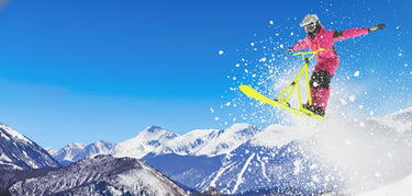 Person on a yellow bike in a pink jacket performing a jump with snowy mountains in the background. cuchara mountain park ski area, affordable skiing, affordable snowboarding, affordable colorado ski areas, affordable colorado ski resort